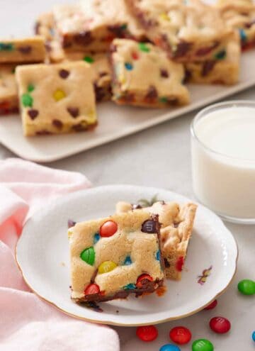 Two pieces of M&M cookie bars on a plate with a platter of more and a glass of milk in the background.