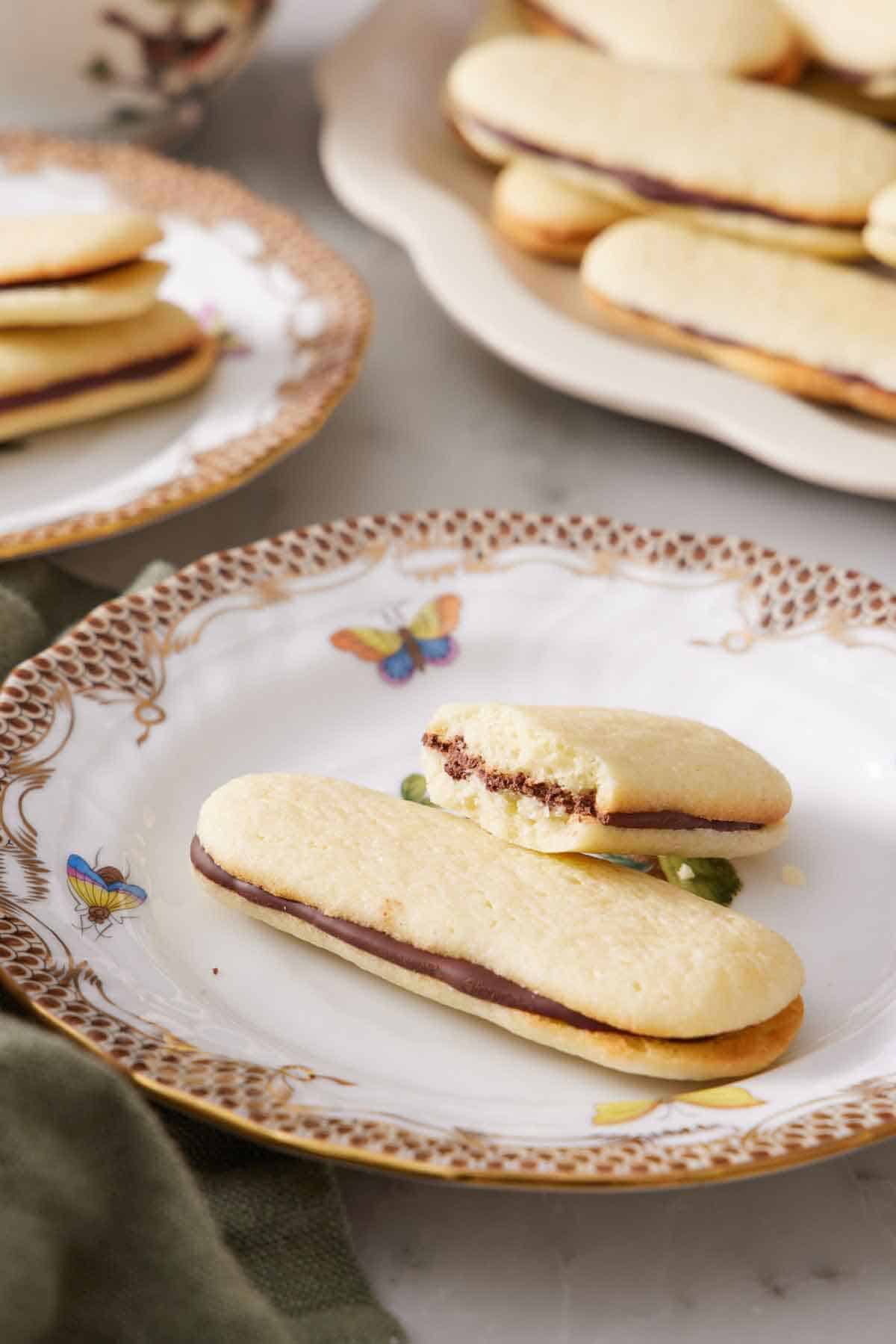 A plate with a Milano cookie and a second half eaten one propped on top. More Milano cookies in the background in a platter and on a plate.