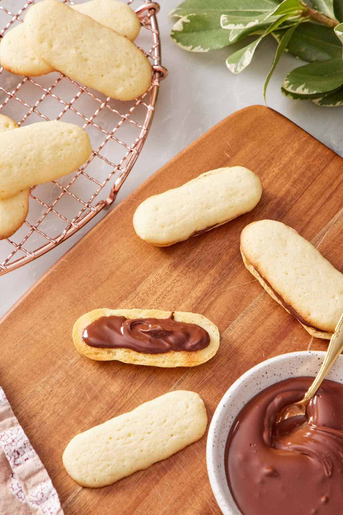 A Milano cookie in the process of being assembled on a wooden serving board with a bowl of melted chocolate on the side and a cooling rack with more cookies.