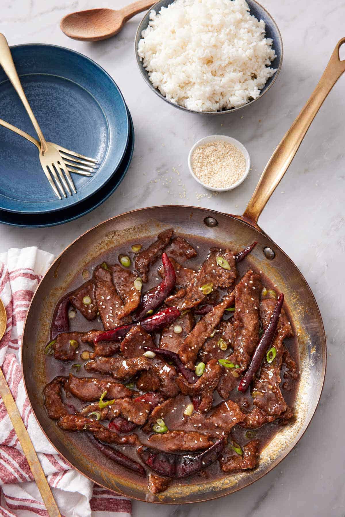 Overhead view of a skillet of Mongolian beef. A bowl of rice, sesame seeds, plates, and forks off to the side.