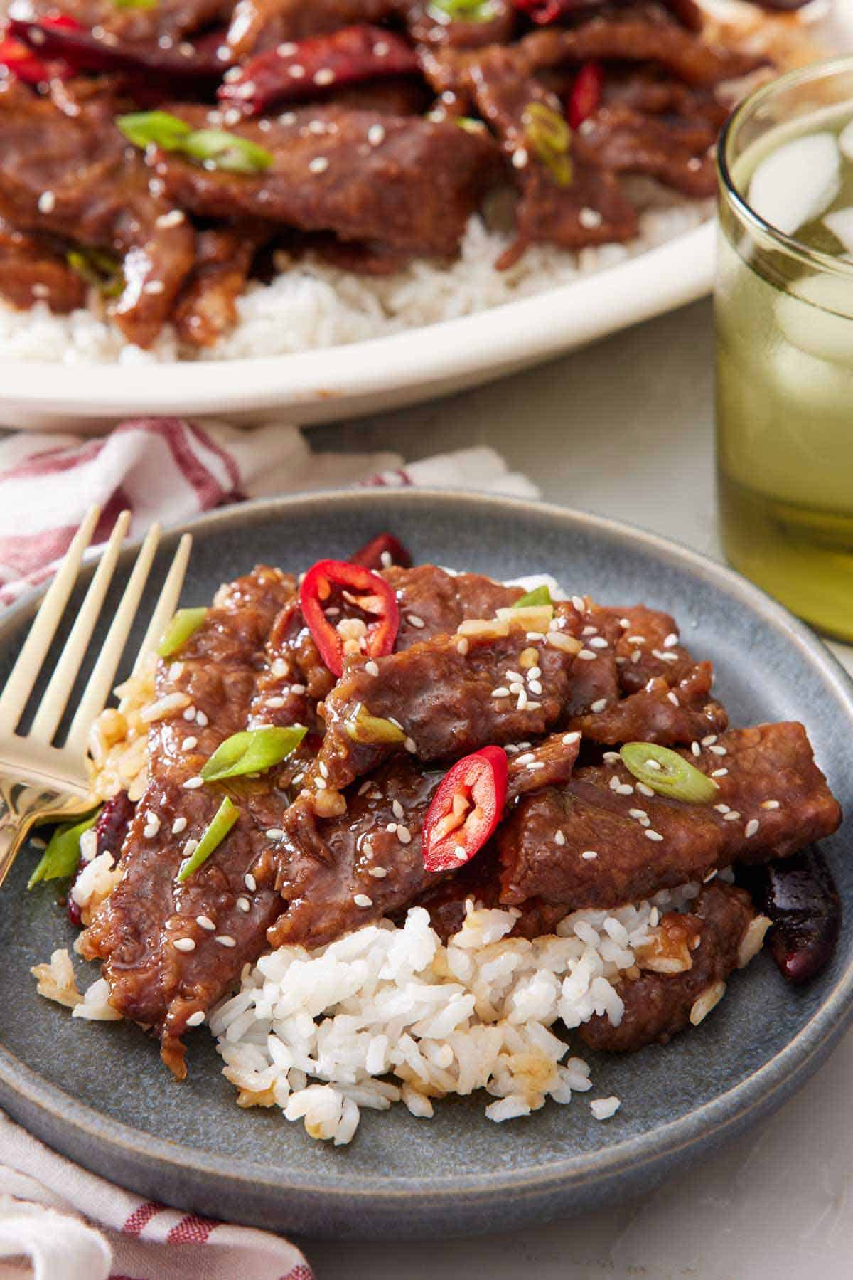 A plate of Mongolian beef over rice, garnished with chili peppers and green onions. A drink and more beef in the background.