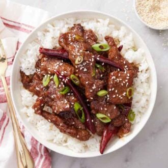 An overhead view of Mongolian beef over rice topped with green onions and sesame seeds. A fork and napkin on the side.