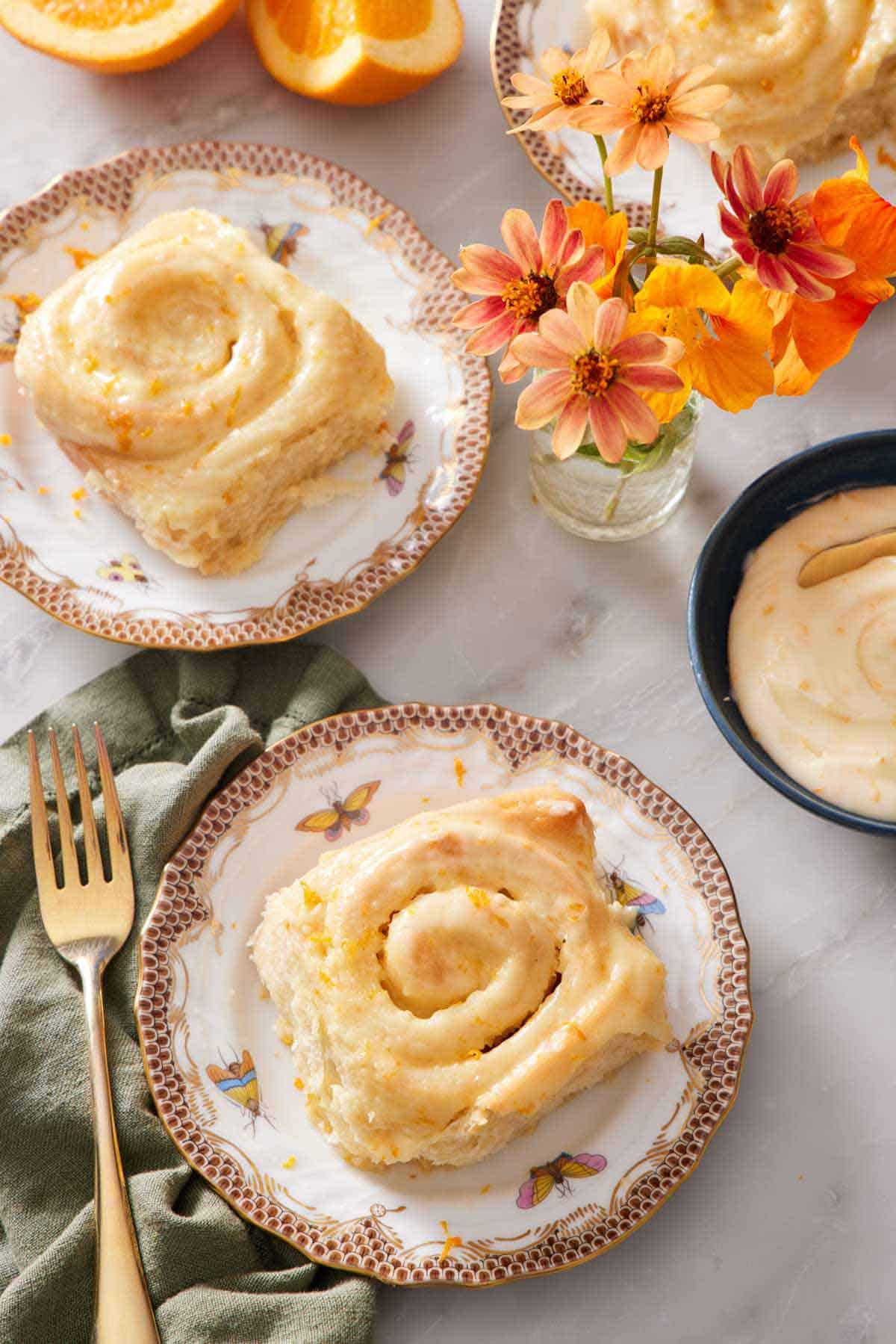 Overhead view of plated orange rolls with a bowl of glaze and orange flowers in a small vase.