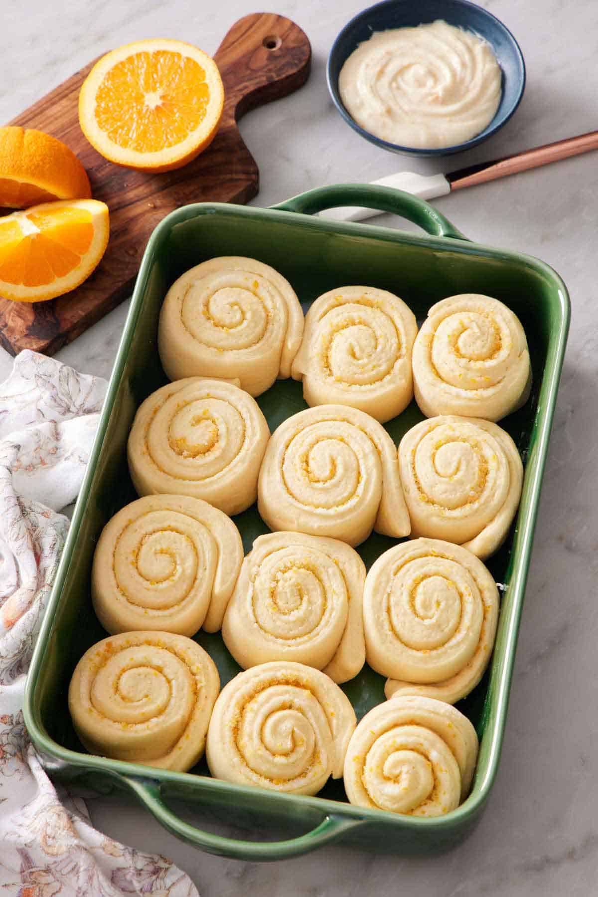 A green baking dish with orange rolls. A bowl of glaze and some cut oranges in the background.