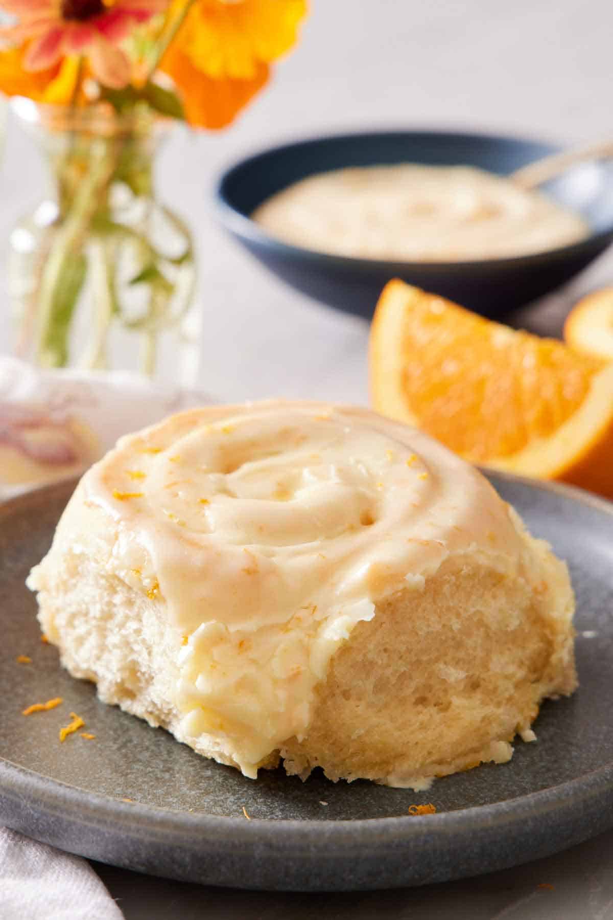 A close up profile view of an orange roll on a plate. Cut orange, bowl of glaze, and orange flowers in the background.