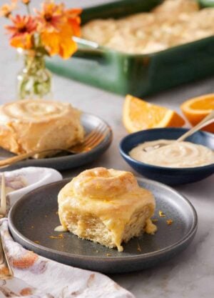 A plate with an orange rolls. A bowl of glaze in the background also with another plated roll and a baking dish with the rest.