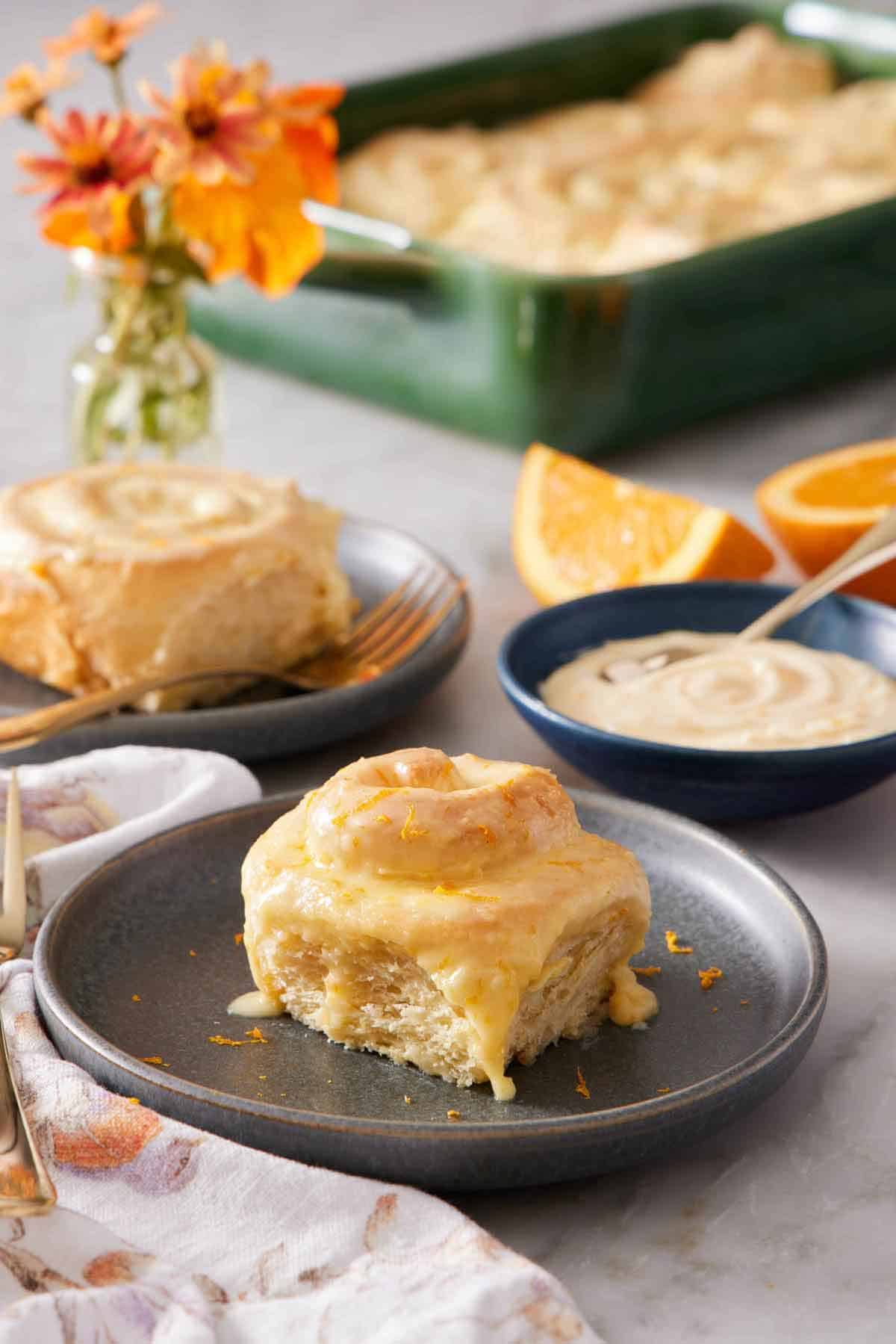 A plate with an orange rolls. A bowl of glaze in the background also with another plated roll and a baking dish with the rest.