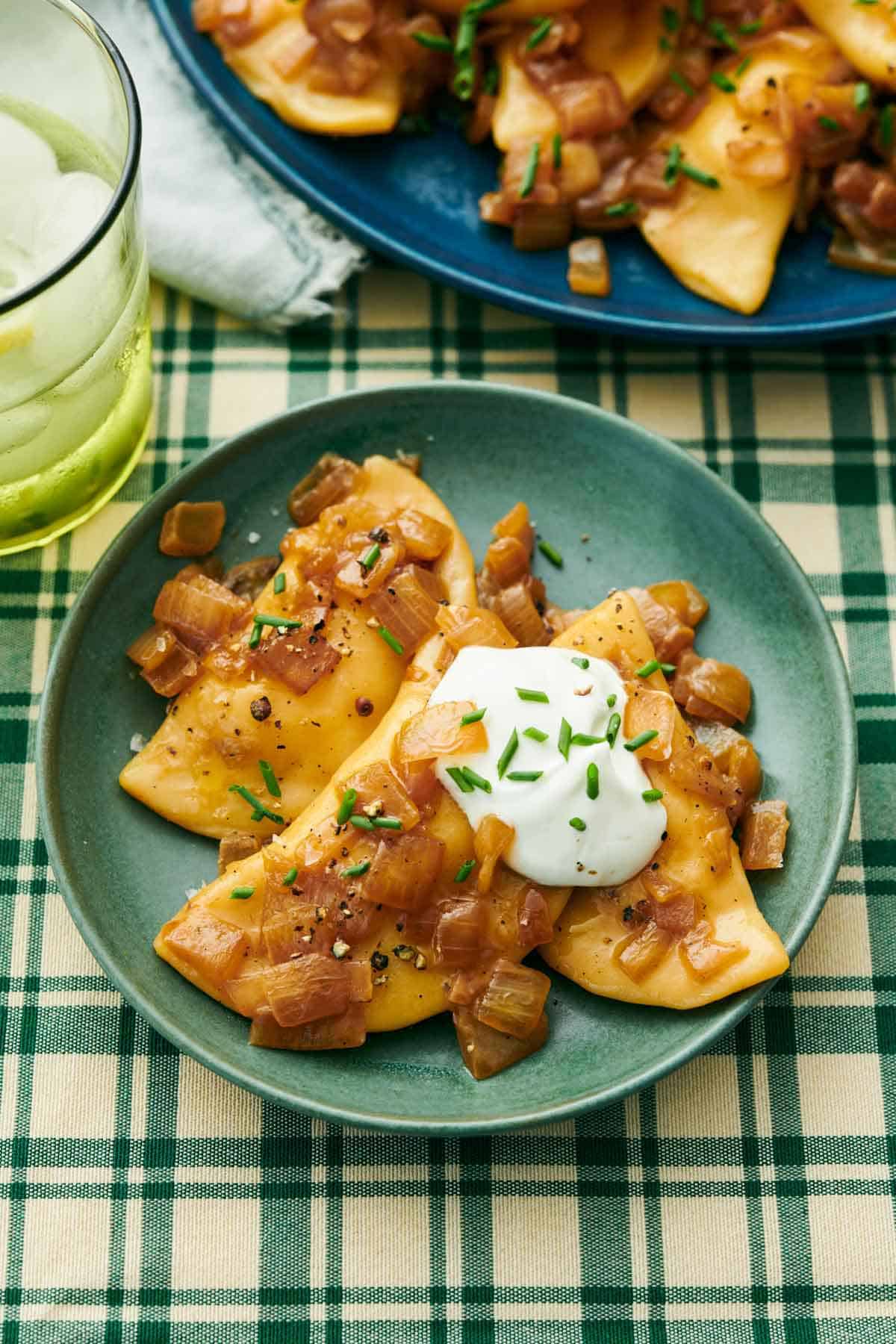 A plate with three pierogies topped with chives and sour cream. A drink and more pierogies in the background.