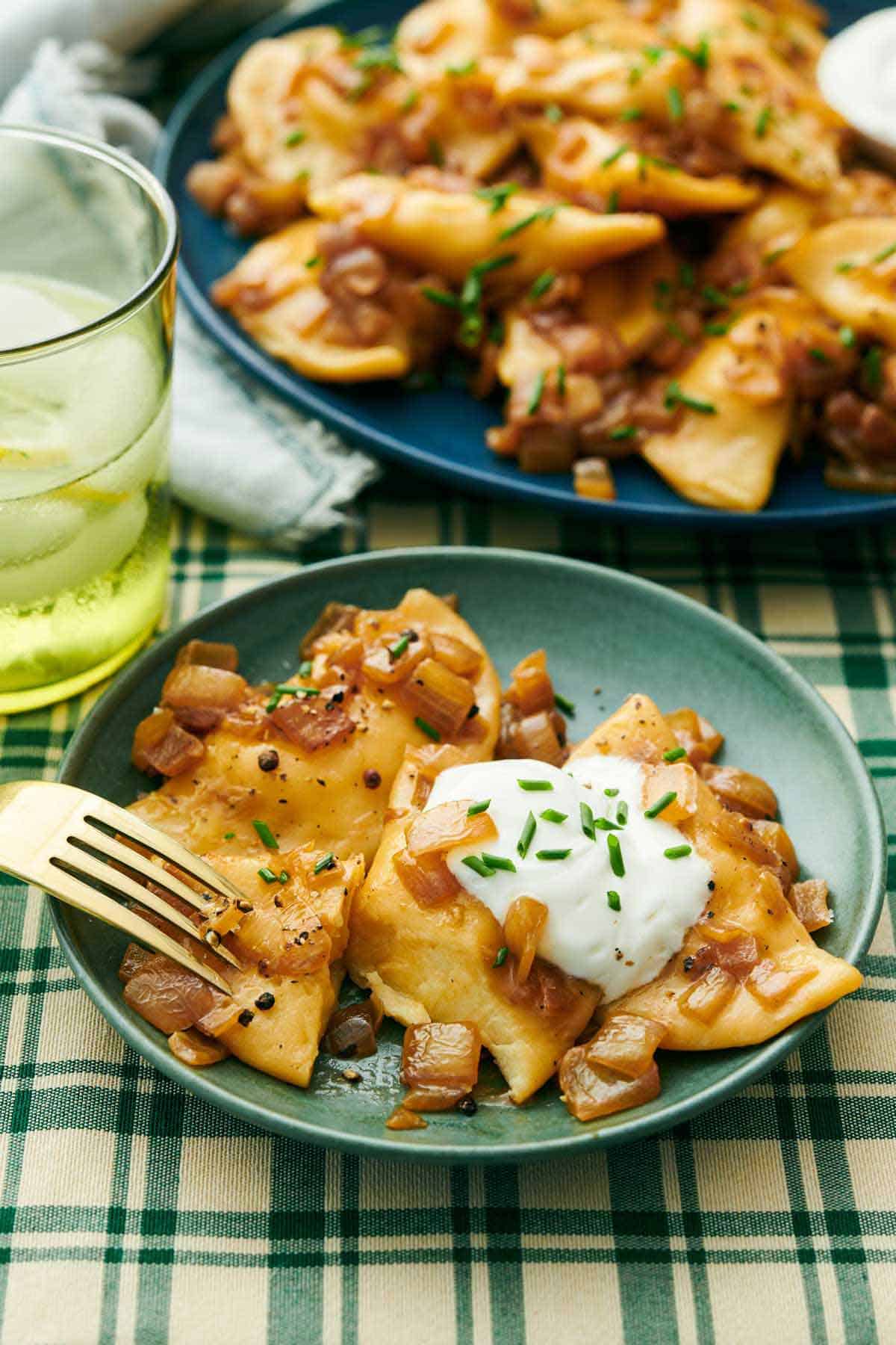 A plate with pierogies, sour cream, and chives with a fork. A platter with more pierogies in the background along with a drink.