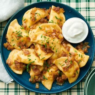 Overhead view of a platter of pierogies with a bowl of sour cream on a checkered tablecloth. Chives garnished on top.