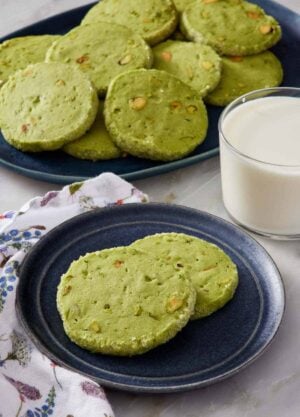 A plate with two pistachio matcha cookies. A glass of milk and platter of more cookies in the background.