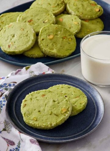 A plate with two pistachio matcha cookies. A glass of milk and platter of more cookies in the background.