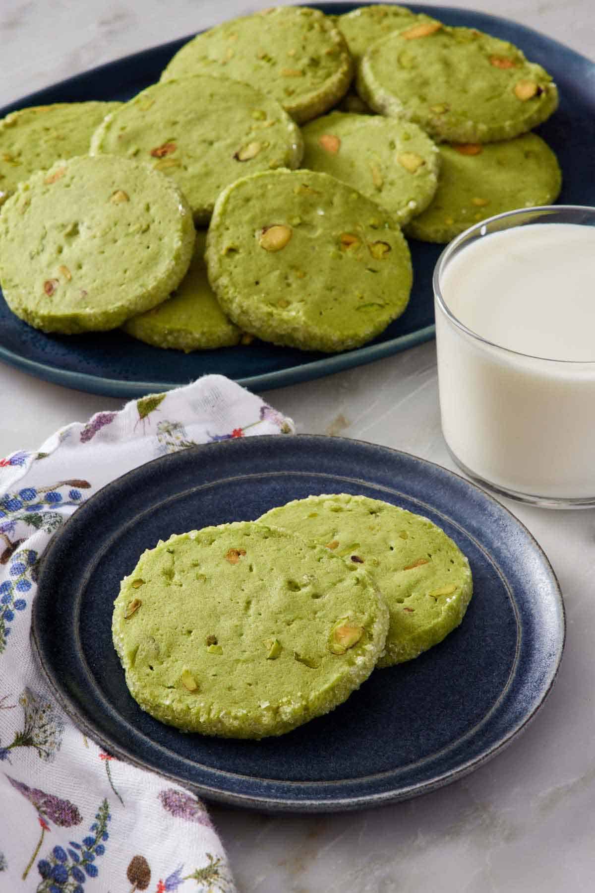 A plate with two pistachio matcha cookies. A glass of milk and platter of more cookies in the background.