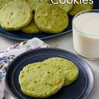 Pinterest graphic of a plate with two pistachio matcha cookies. A glass of milk and platter of more cookies in the back.