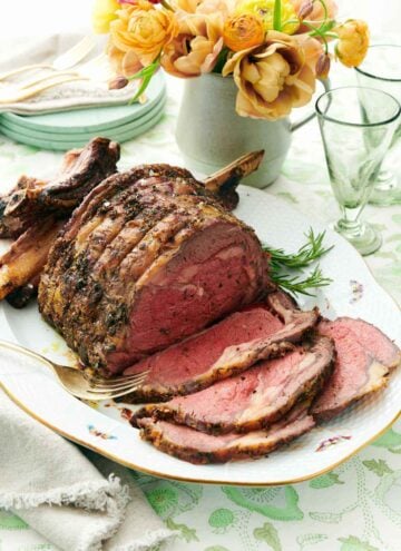 A platter with a prime rib with a few slices cut in front of it. A vase of flowers in the back along with some cups and plates.