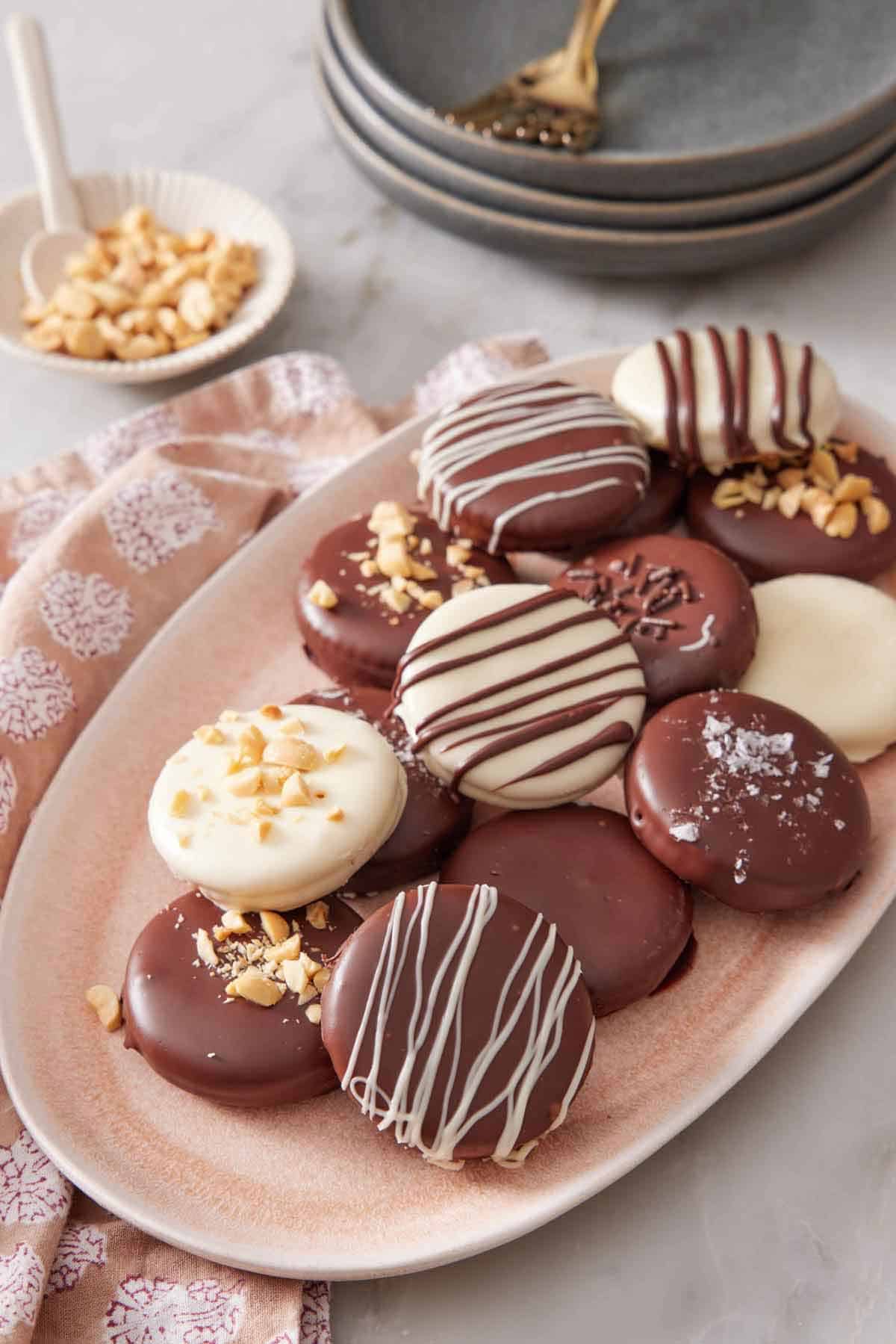 A platter of variously decorated Ritz cookies. A bowl of chopped peanuts in the background along with a stack of plates and forks.