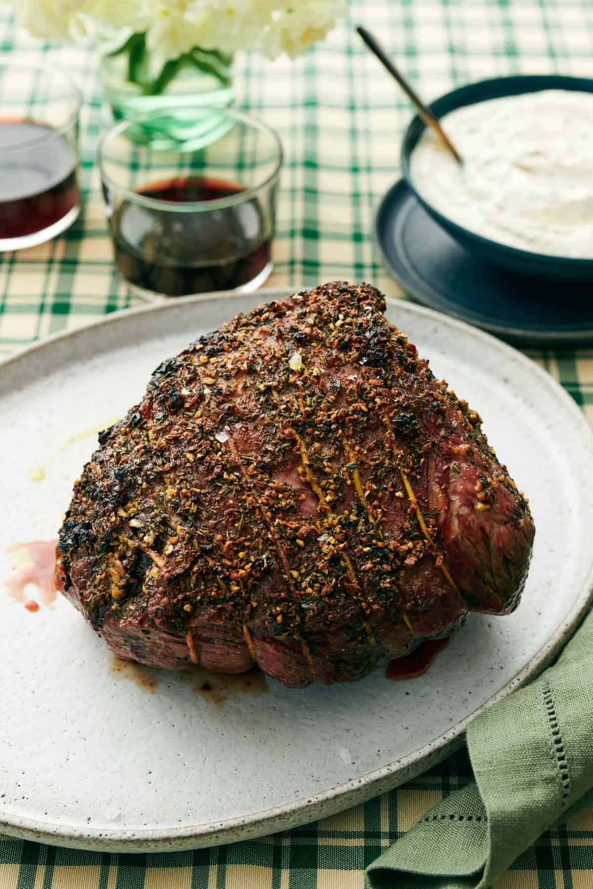 A roast beef on an oval platter with a bowl of horseradish sauce in the back along with two drinks.