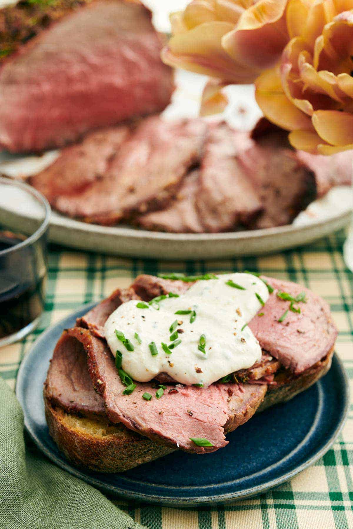 A plate with a thick piece of bread topped with sliced roast beef and horseradish sauce with chives. More sliced roast beef in the background.