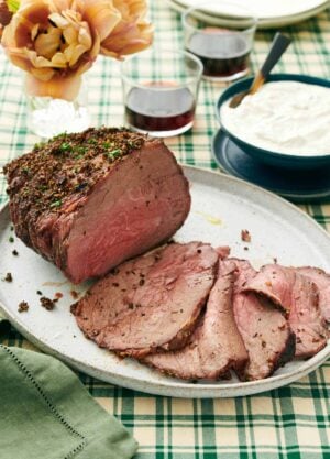 A platter with a roast beef with half of it sliced in front. Sauce in the background along with drinks and flowers.