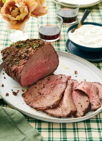 A platter with a roast beef with half of it sliced in front. Sauce in the background along with drinks and flowers.