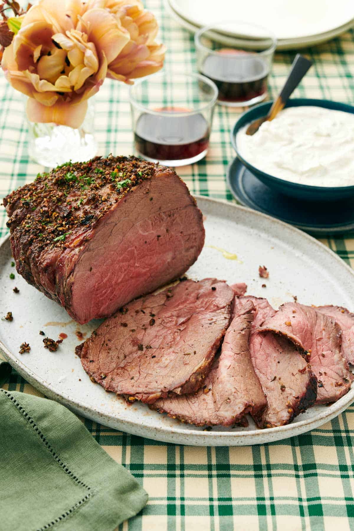 A platter with a roast beef with half of it sliced in front. Sauce in the background along with drinks and flowers.