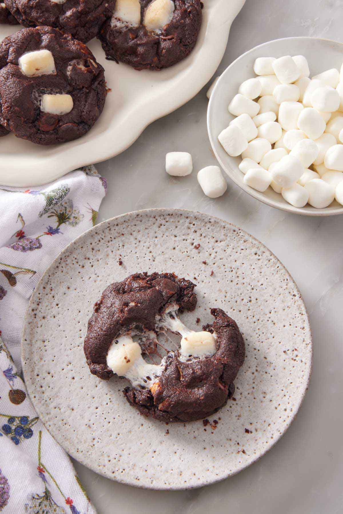 A plate with a rocky road cookie torn apart. A bowl of mini marshmallows and more cookies in the background.