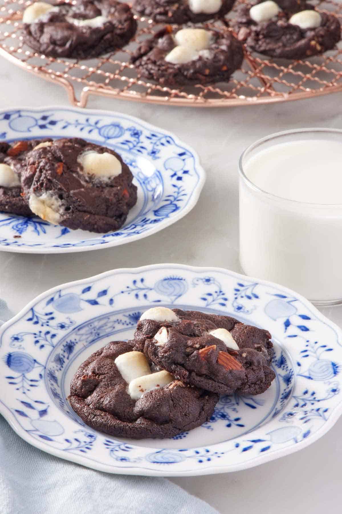 A plate with two rocky road cookies with a glass of milk, another plate of cookies, and a cooling rack of cookies in the background.