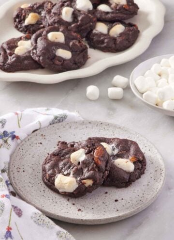 A plate with two rocky road cookies and a platter more in the background. A bowl of mini marshmallows on the side.