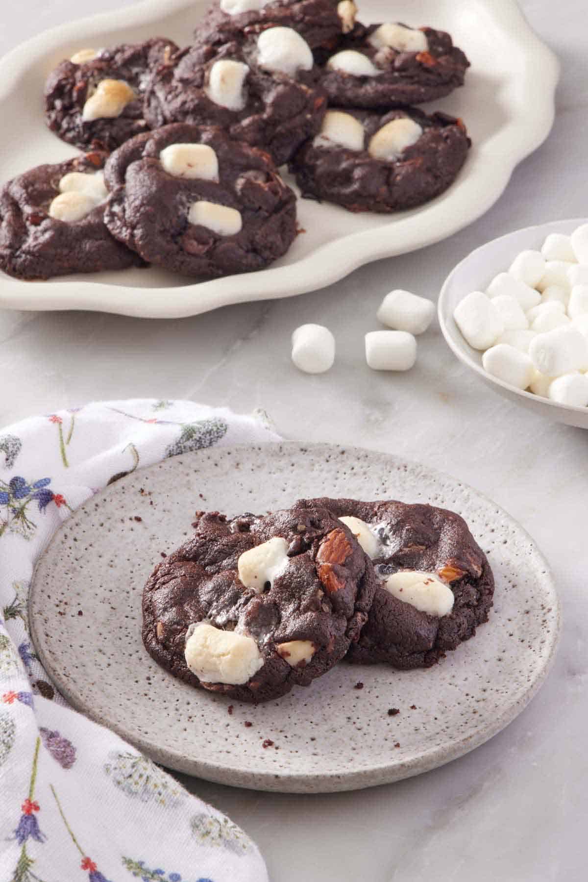 A plate with two rocky road cookies and a platter more in the background. A bowl of mini marshmallows on the side.