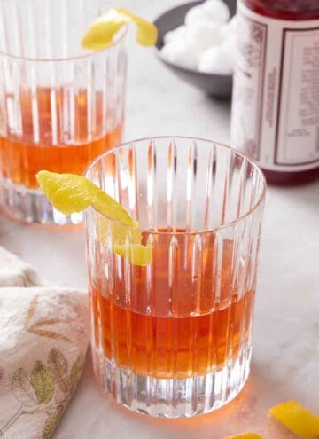 Two glasses of Sazerac with a lemon twist garnish on the rims of the glasses. Sugar cubes and a bottle of bitters in the back.