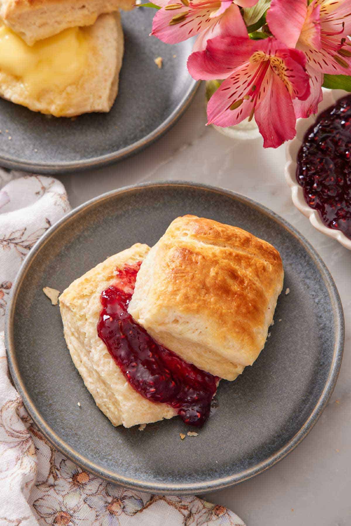 A plate with a sourdough biscuit cut in half with jam in between. Flowers, jam, and another biscuit in the background.