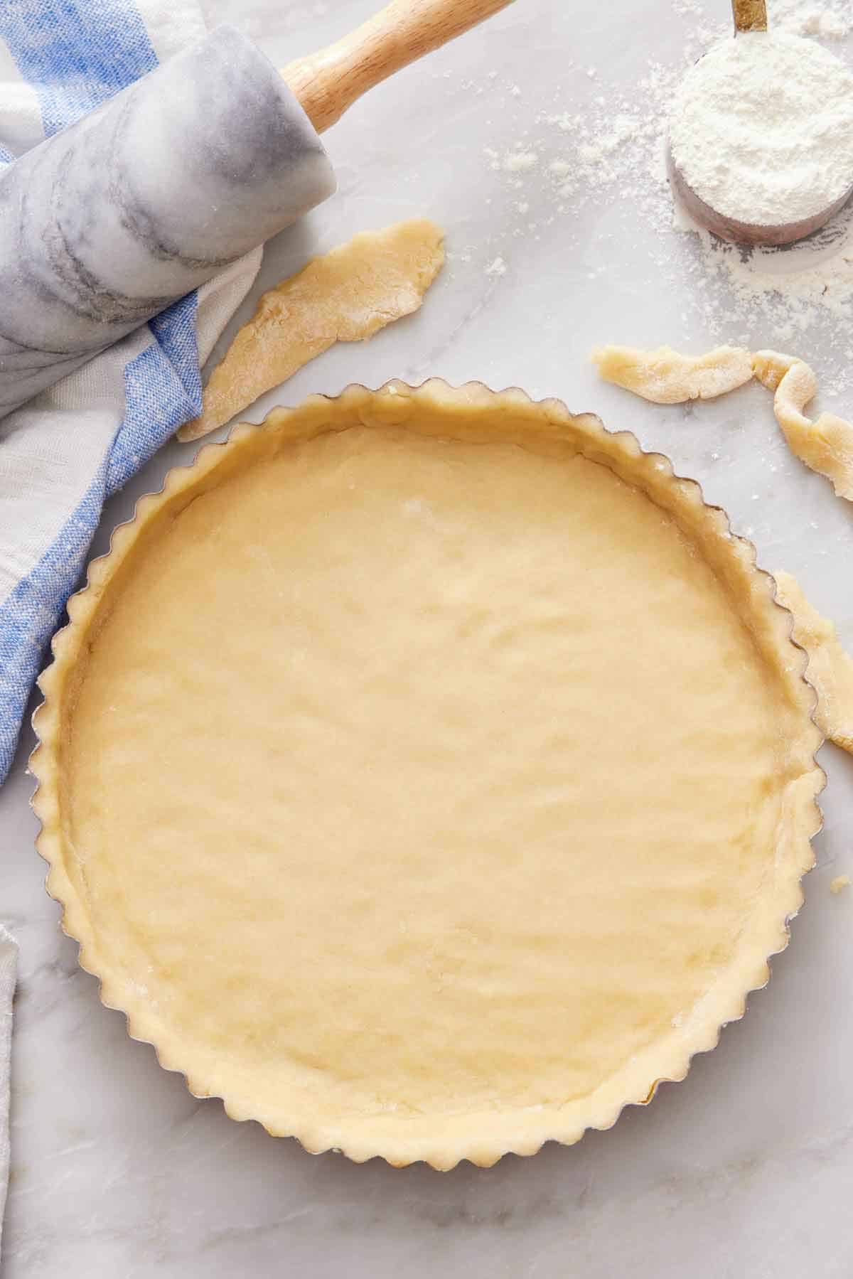 An overhead view of a tart crust before baking. A rolling pin and cup of flour in the background.