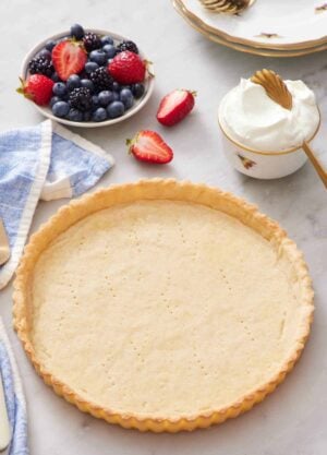 A baked tart crust with a bowl of berries and whipped cream in the background.