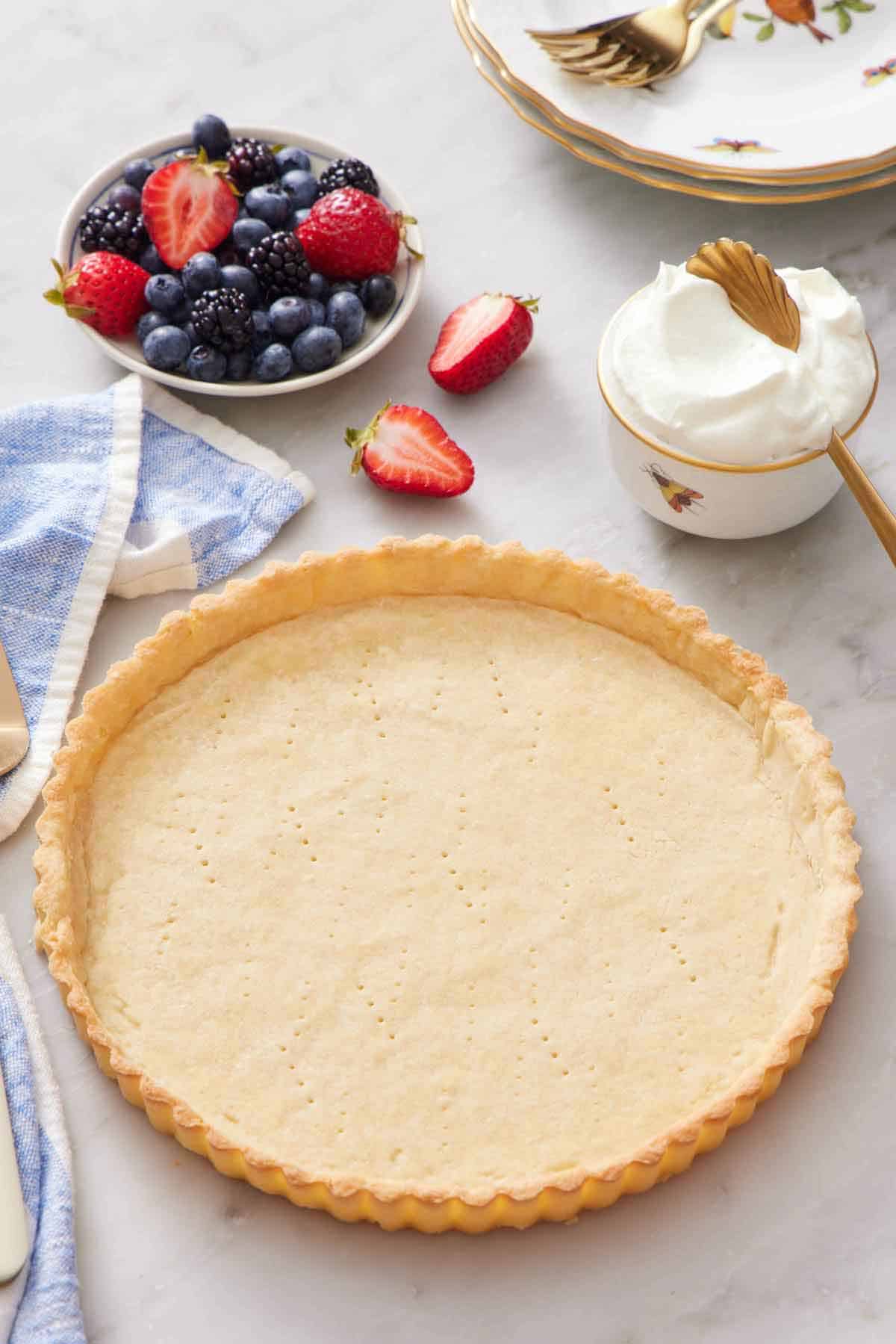 A baked tart crust with a bowl of berries and whipped cream in the background.