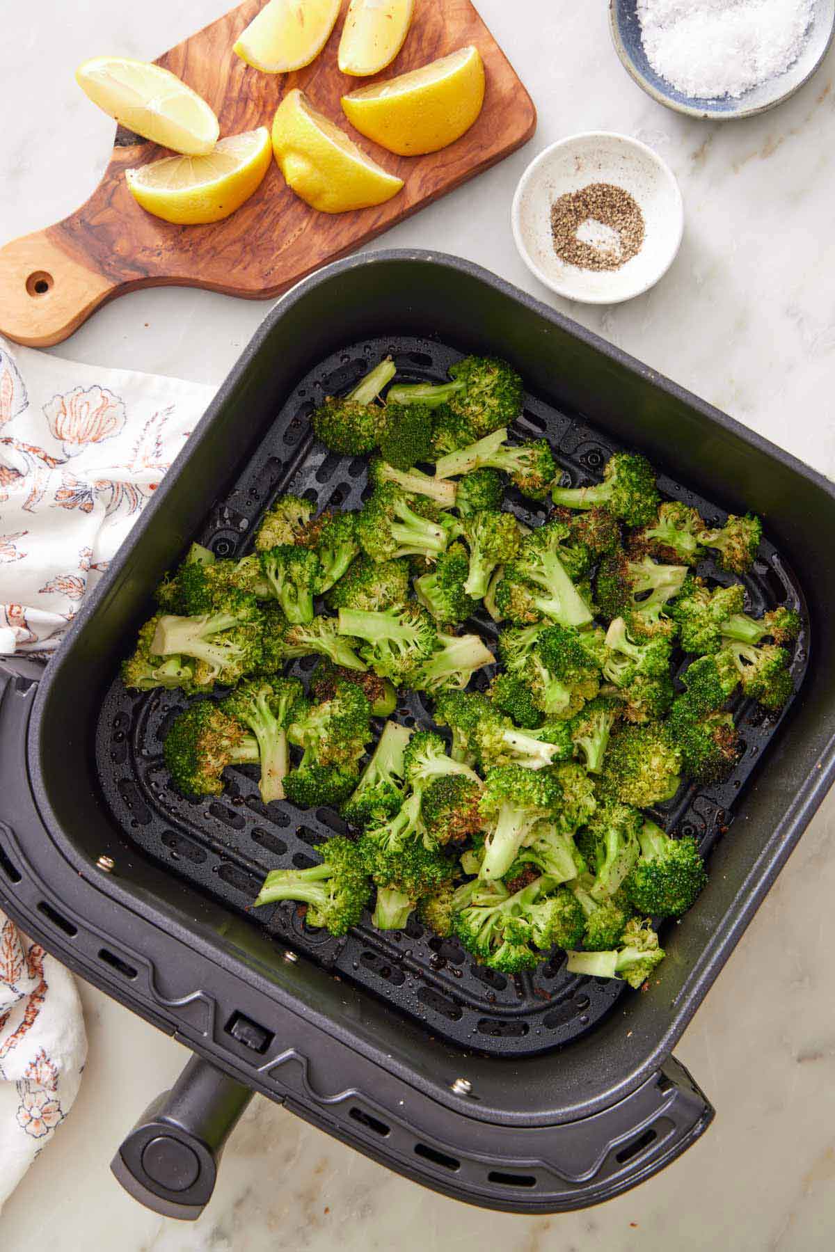 Overhead view of air fryer broccoli in an air fryer basket. Cut lemon wedges, a bowl of pepper, and bowl of salt on the side.