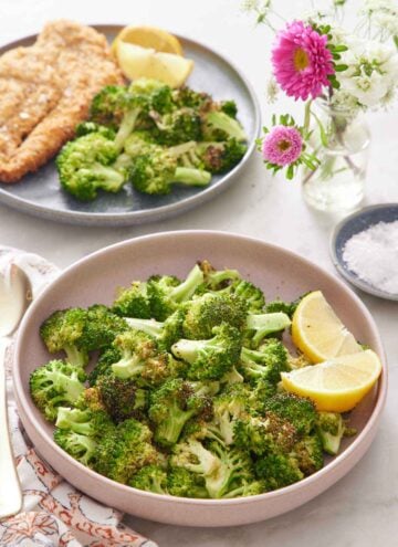 A pink plate of air fryer broccoli with lemon wedges. A vase of flowers in the back along with a plate of fried cutlet, broccoli, and lemon wedges. A bowl of salt on the side.