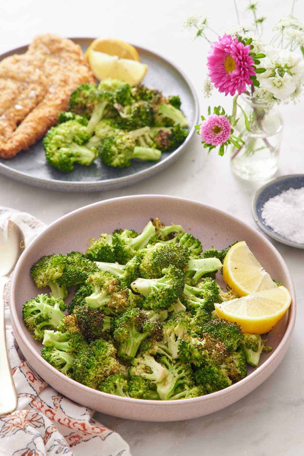 A pink plate of air fryer broccoli with lemon wedges. A vase of flowers in the back along with a plate of fried cutlet, broccoli, and lemon wedges. A bowl of salt on the side.