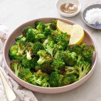 A plate of air fryer broccoli with two lemon wedges. A linen napkin with a serving fork on the side along with salt and pepper in bowls.