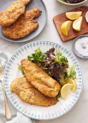 A plate with crispy air fryer chicken cutlets along with a side of mixed greens and lemon wedges. More cutlets in the back along with cut lemon wedges and a bowl of salt.