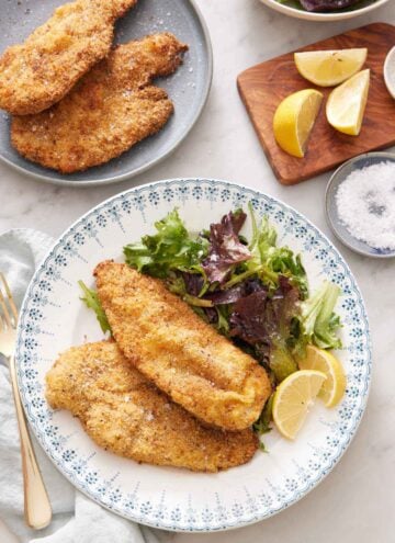 A plate with crispy air fryer chicken cutlets along with a side of mixed greens and lemon wedges. More cutlets in the back along with cut lemon wedges and a bowl of salt.