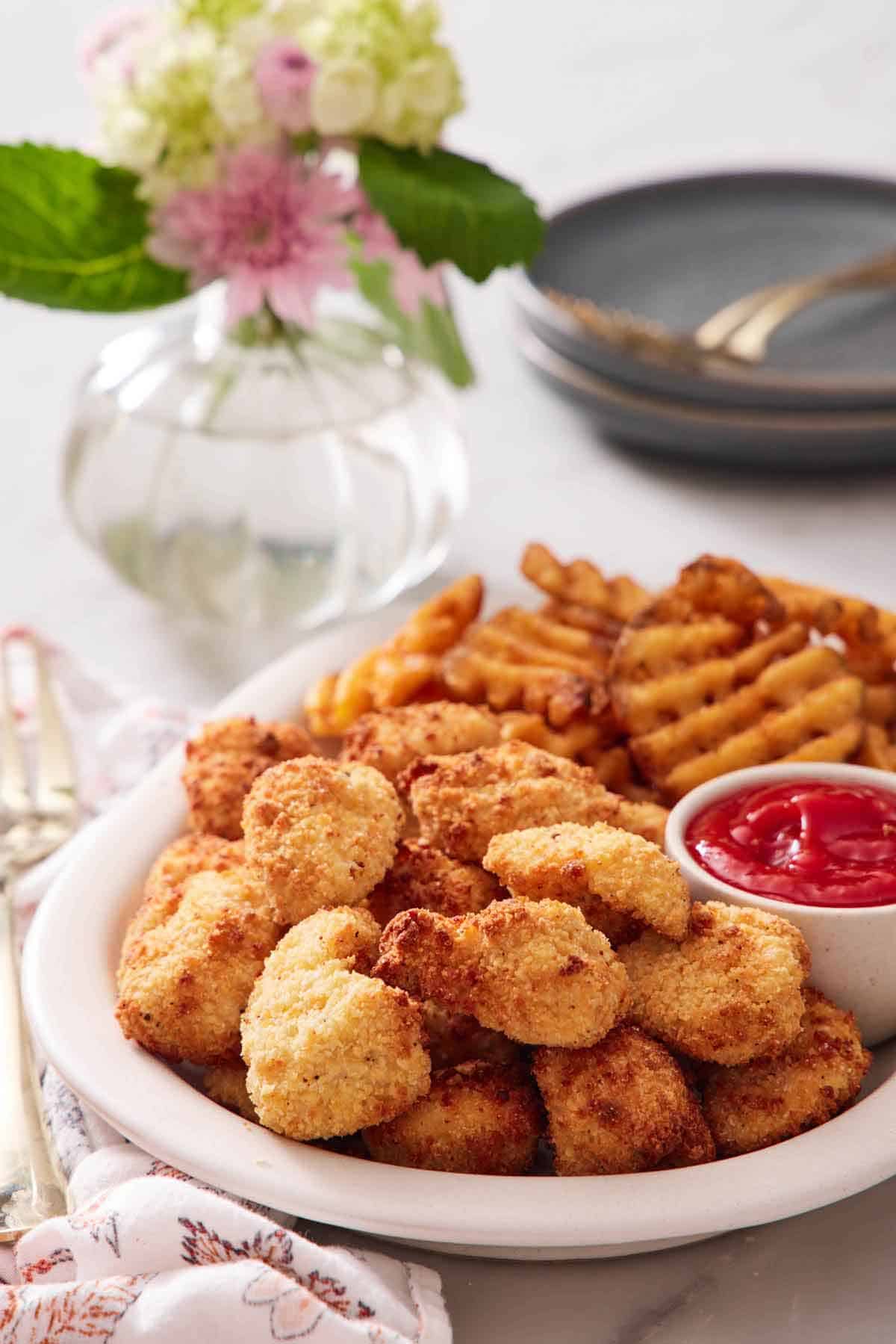 A platter with air fryer chicken nuggets, ketchup, and waffle fries. A small vase of flowers in the back.