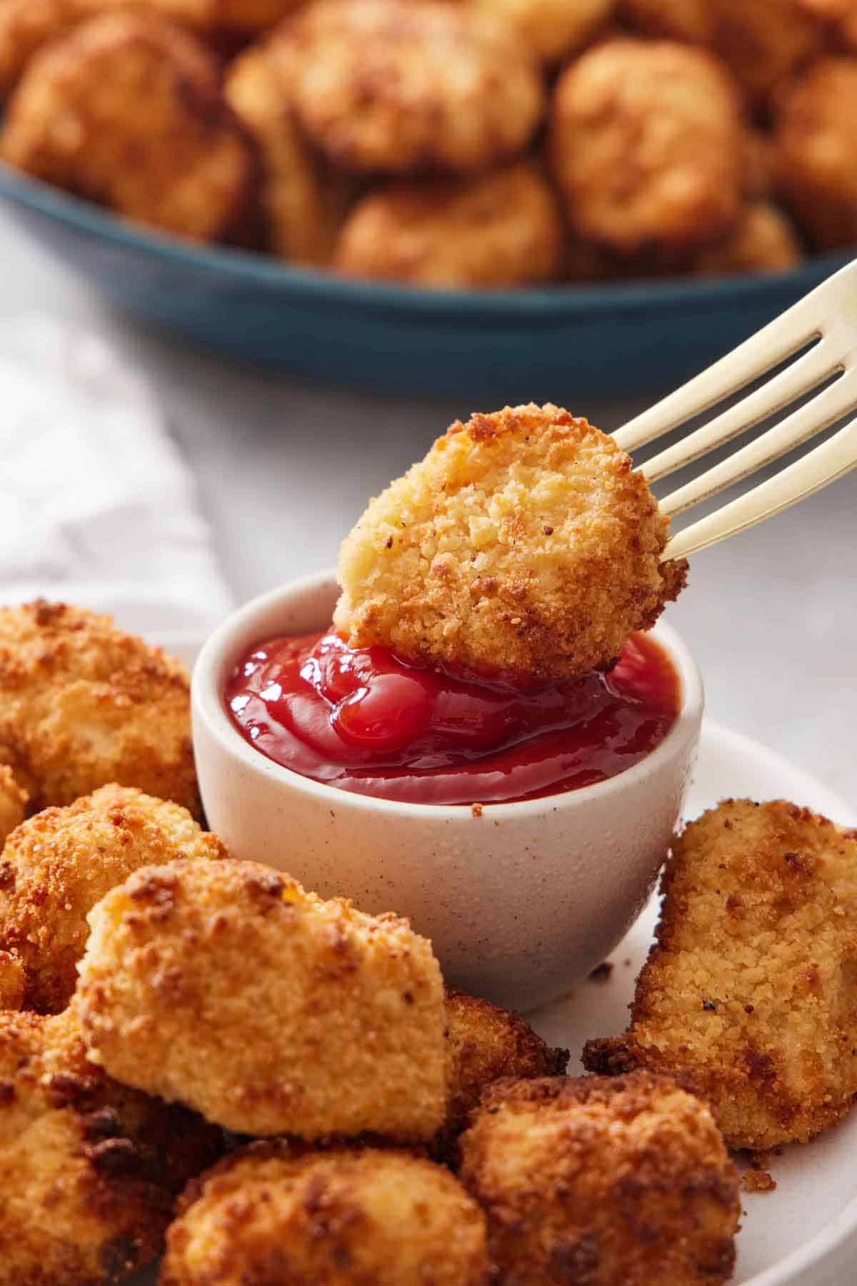 An air fryer chicken nugget on a fork dipped into a small bowl of ketchup. More air fryer chicken nuggets in the background.