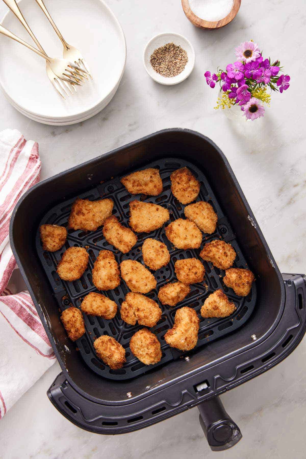 Overhead view of air fryer chicken nuggets in an air fryer basket. Flowers, plates, forks, salt, and pepper in the background.