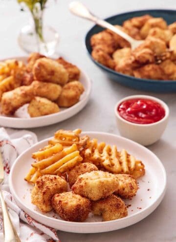 A plate with air fryer chicken nuggets and waffle fries. A bowl of ketchup in the back along with more air fryer chicken nuggets and waffle fries.