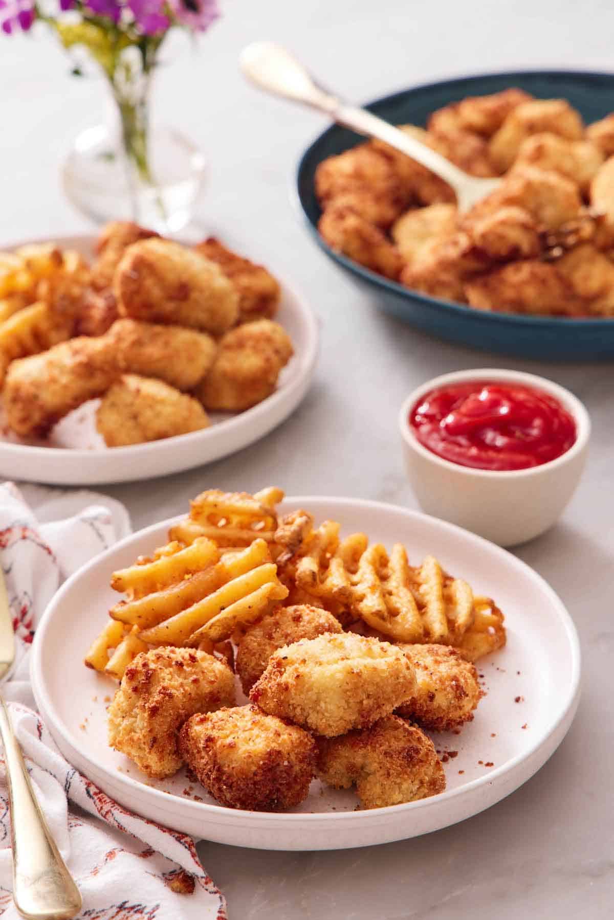 A plate with air fryer chicken nuggets and waffle fries. A bowl of ketchup in the back along with more air fryer chicken nuggets and waffle fries.