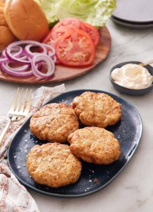 A platter with four air fryer chicken patties. Burger toppings in the back along with a bowl of sauce.