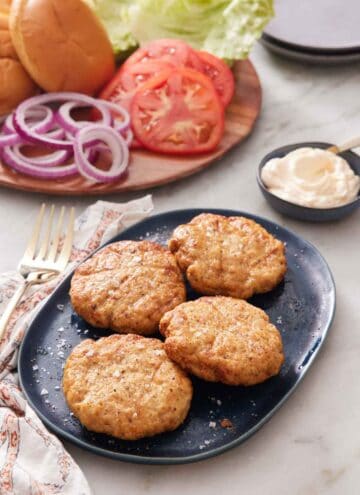 A platter with four air fryer chicken patties. Burger toppings in the back along with a bowl of sauce.