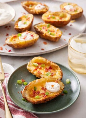 A plate with two air fryer potato skins with a platter in the background with more air fryer potato skins. A drink on the side.