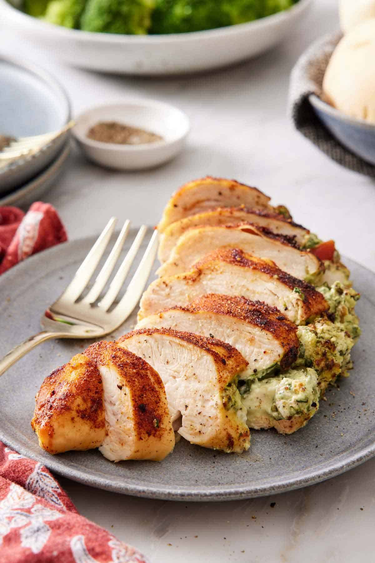 An air fryer stuffed chicken breast on a plate, sliced. A fork beside the chicken. Some pepper in the background along with broccoli and bread.