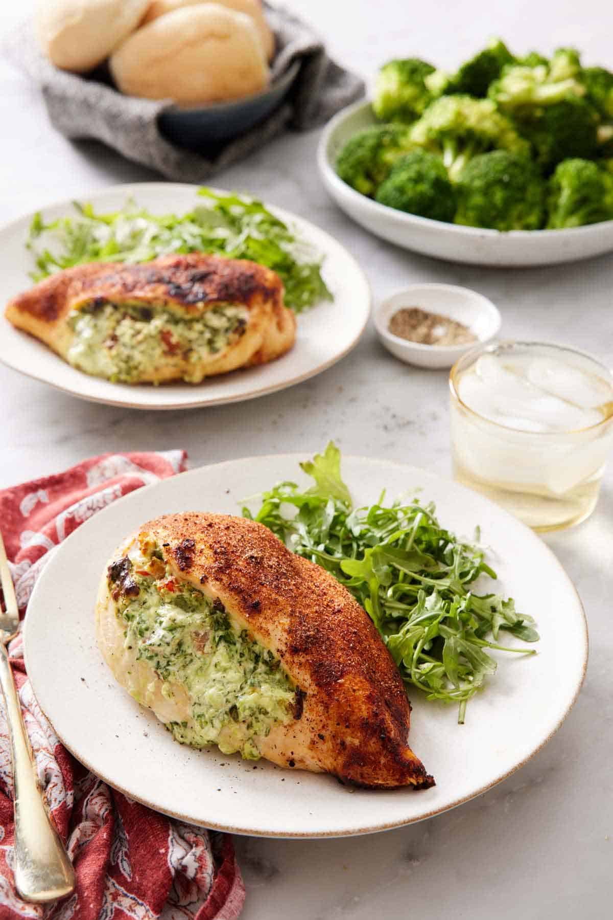 A plate with an air fryer stuffed chicken breast with some arugula. A drink, another plated serving, broccoli, and bread in the background.